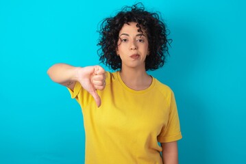 young arab woman wearing yellow T-shirt over blue background looking unhappy and angry showing...