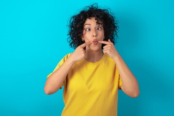 young arab woman wearing yellow T-shirt over blue background crosses eyes and makes fish lips funny grimace