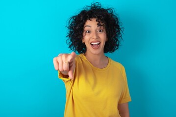 Excited positive young arab woman wearing yellow T-shirt over blue background points index finger...