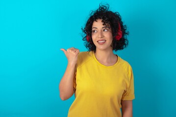 young arab woman wearing yellow T-shirt over blue background listens audio track via wireless headphones points thumb away advertises copy space