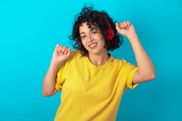 Carefree young arab woman wearing yellow T-shirt over blue background with toothy smile raises arms dances carefree moves with rhythm of music listens music from playlist via headphones