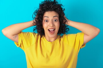 Cheerful overjoyed young arab woman wearing yellow T-shirt over blue background reacts rising hands over head after receiving great news.