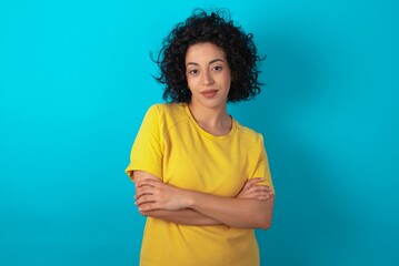 Confident young arab woman wearing yellow T-shirt over blue background with arms crossed looking to the camera