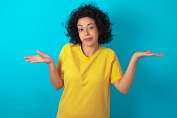 Careless attractive young arab woman wearing yellow T-shirt over blue background shrugging...