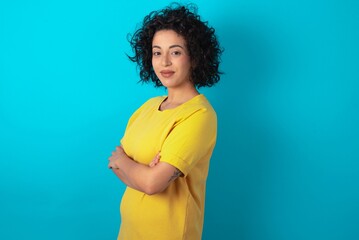 Portrait of young arab woman wearing yellow T-shirt over blue background standing with folded arms...