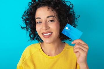 Close up photo of optimistic young arab woman wearing yellow T-shirt over blue background hold card