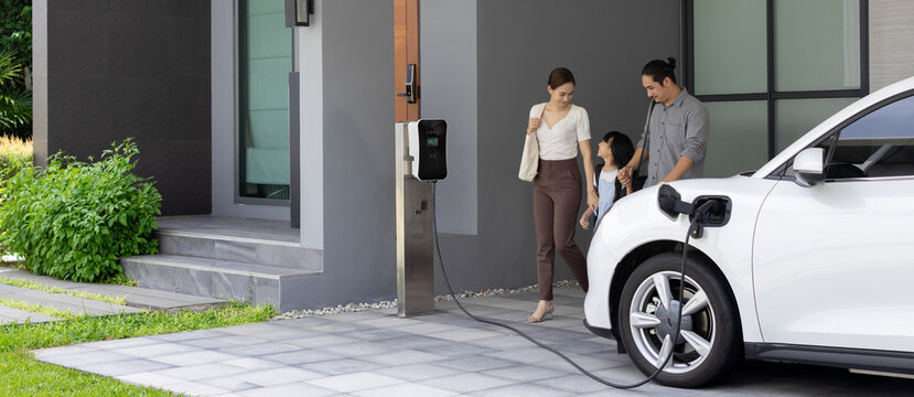 Naklejka Progressive young parents and daughter with electric vehicle and home charging station. Green and clean energy from electric vehicles for healthy environment. Eco power from renewable source at home.