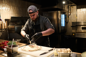 Young chef in uniform and black protective gloves sprinkling grated cheese on flatbread while cooking pizza in the kitchen of luxurious restaurant