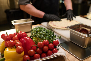 Pile of fresh and ripe vegetables prepared for cooking pizza in metallic tray on table against chef in gloves putting ingredients on flatbread