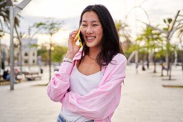 Fototapeta premium Cheerful and pretty Chinese woman using smartphone being on a call while standing outdoors during a summer day.