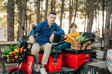 Fototapeta na wymiar Farmer father and school boy kid delivered wooden crates with vegetables to camp site for picnic. Dad man male and son tourists delivered groceries on electric tricycle vehicle in the forrest