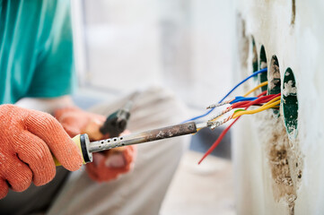 Close up of man in work gloves using electric repair solder welding tool while installing electrical cables in apartment under renovation. Male electrician working with electric heater soldering iron.