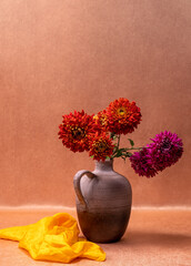 A bouquet of red, pink dahlias in a vase on an orange background, next to it lies a yellow scarf. Still life, postcard