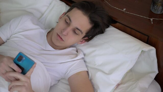 Top view portrait of young man scrolling social media in phone application lying in soft comfortable bed. Sleepless Caucasian guy procrastinating at bedtime at home indoors