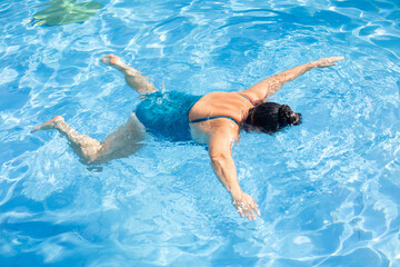 Portrait of tanned middle-aged woman wearing blue swimsuit, floating on stomach in swimming pool, relaxing on sunny day.