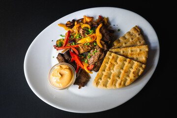 Top view - Beef fajitas served with pita bread and spicy dip on white plate and black background