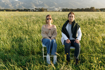 Fototapeta na wymiar Young happy couple sitting on chairs in feather grass field on summer sunset day, looking forward in camera, serious