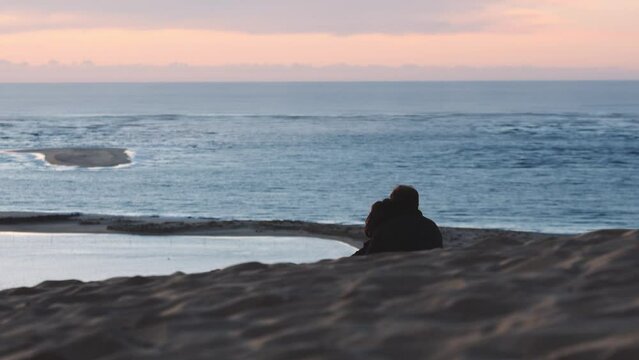 Romantic couple sitting alone on sand Dune of Pilat hugging watching sunset