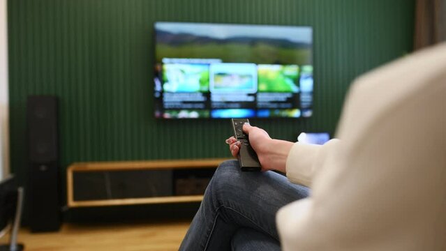 Woman is sitting at home watching TV, holding remote controller.