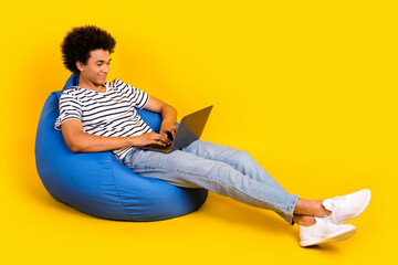 Full body size photo of young satisfied guy lying beanbag with netbook writing his graduation last exam isolated on yellow color background