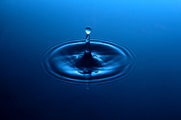 A brilliant rich blue pool of water gently rippled with concentric circles with a single drop of connected spherical water splash above a column of liquid.