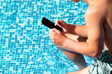Young man using smartphone in resort hotel poolside during summer holidays, vacation. Guy hold mobile phone isolated on blue water swimming pool background copy space. Remote work, online banking