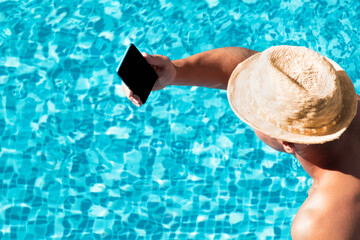 Young man using smartphone in resort hotel poolside during summer holidays, vacation. Guy in straw hat hold mobile phone isolated on blue water swimming pool background copy space