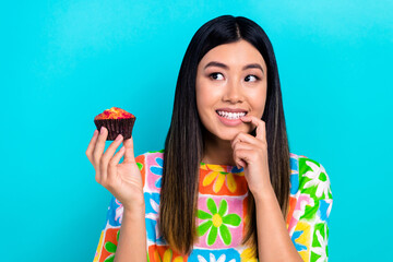 Photo of shiny doubtful woman dressed flower print t-shirt eating tasty cupcake looking empty space...