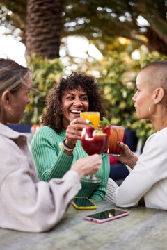 Three cheerful mature women having drinks outdoors in a terrace, toasting with cocktails or mocktails. Female friends having fun together gathering in a bar. Vertical image.