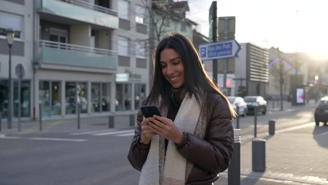One happy young woman looking at cellphone device standing in city street during sunset time. 20s female adult girl holding phone and smiling in urban setting