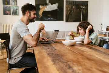 Father working on laptop while his son eating flakes sitting at table at home