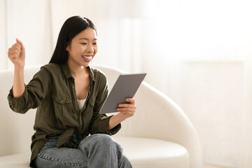 Obraz premium Emotional asian lady sitting on couch with tablet, copy space