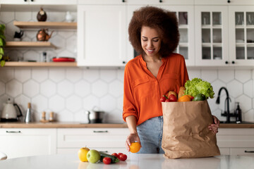 Happy Young Black Lady Unpacking Paper Bag With Groceries After Food Shopping