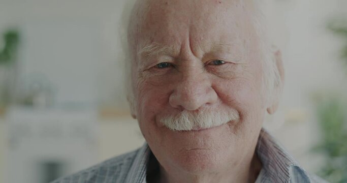 Close-up portrait of cheerful elderly man smiling looking at camera indoors in apartment. Face expression and happy retirement concept.