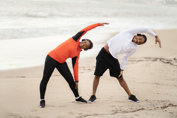 Glad young african american woman and man in sportswear and headphones doing stretching exercise