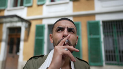 One pensive serious young Middle Eastern man smoking a cigarette outside with thoughtful expression. Tracking shot closeup face in motion while brooding