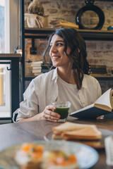 Fototapeta premium A young attractive dark-haired woman is reading a book in a cafe and drinking green matcha tea.