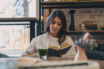 A young attractive dark-haired woman is reading a book in a cafe and drinking green matcha tea.