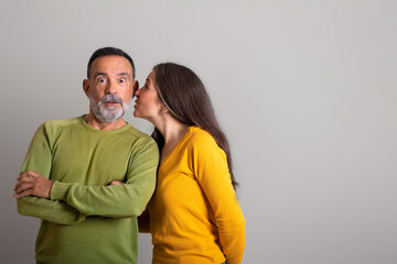 Smiling caucasian elderly wife whispering secret in ear of surprised man on gray studio background