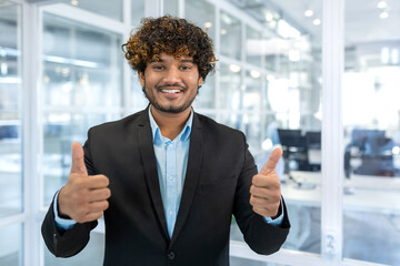 Fototapeta premium Portrait of successful hispanic businessman boss, manager in business suit and beard looking at camera and smiling standing near window, showing thumbs up, sign of success and achieving goals.