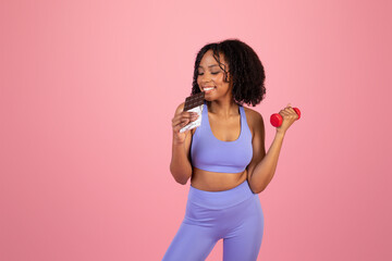 Cheerful millennial african american woman in sportswear with dumbbells eating chocolate