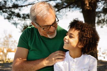 Cheerful senior caucasian grandfather hugging little mixed race boy, talking, enjoy free time, fun outdoor, close up