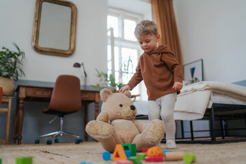 Little boy playing with bear toy and cubes at home.
