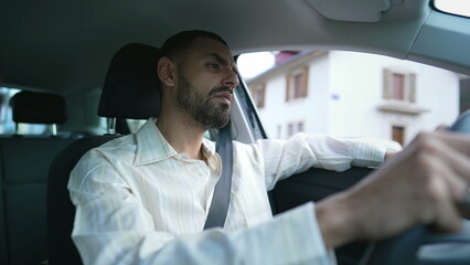 Naklejka na ściany i meble A focused Middle Eastern driver behind the wheel with a pensive expression. An Arab man driving car with care and attention. Interior shot of a young man holding steering wheel with a thoughtful look
