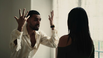 Angry man arguing with girlfriend by window at home. Male person raising arms in frustration and...