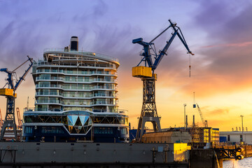 Scenic back view large modern luxury cruise ship liner under construction building cranes at dry dock shipyard in Hamburg port dramatic sunset sky background. Big vessel manufactoring site industry - obrazy, fototapety, plakaty