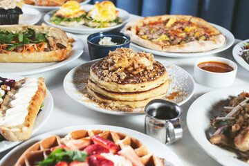 A view of a table full of a variety of food entrees, featuring pancakes, pizza, waffles, and a sandwich.