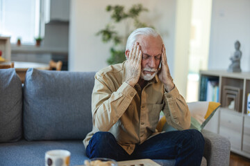 Depressed senior man sitting in the living room. Frustrated senior man with headache covering face and contemplating.