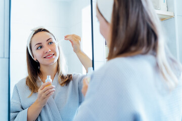 Young woman in bathrobe looking in the mirror, holding dropper with hyaluronic acid serum close to face and smiling. Skin hydrating. Cosmetic spa procedures. Beauty self-care at home. Selective focus.