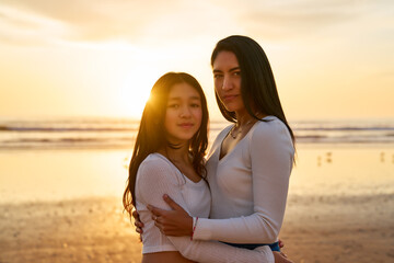 waist up portrait latina mother and daughter embraced on the beach at sunset	